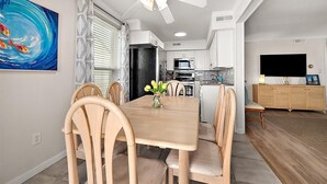Een eethoek met een houten tafel, stoelen, een plafondventilator, een keuken op de achtergrond en een televisie op een kast.