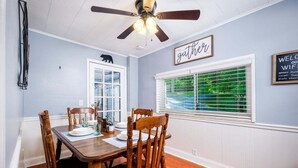 Un coin repas avec une table et des chaises en bois, un ventilateur de plafond, une fenêtre avec des stores et un panneau mural sur lequel est écrit le mot «rassemblement».
