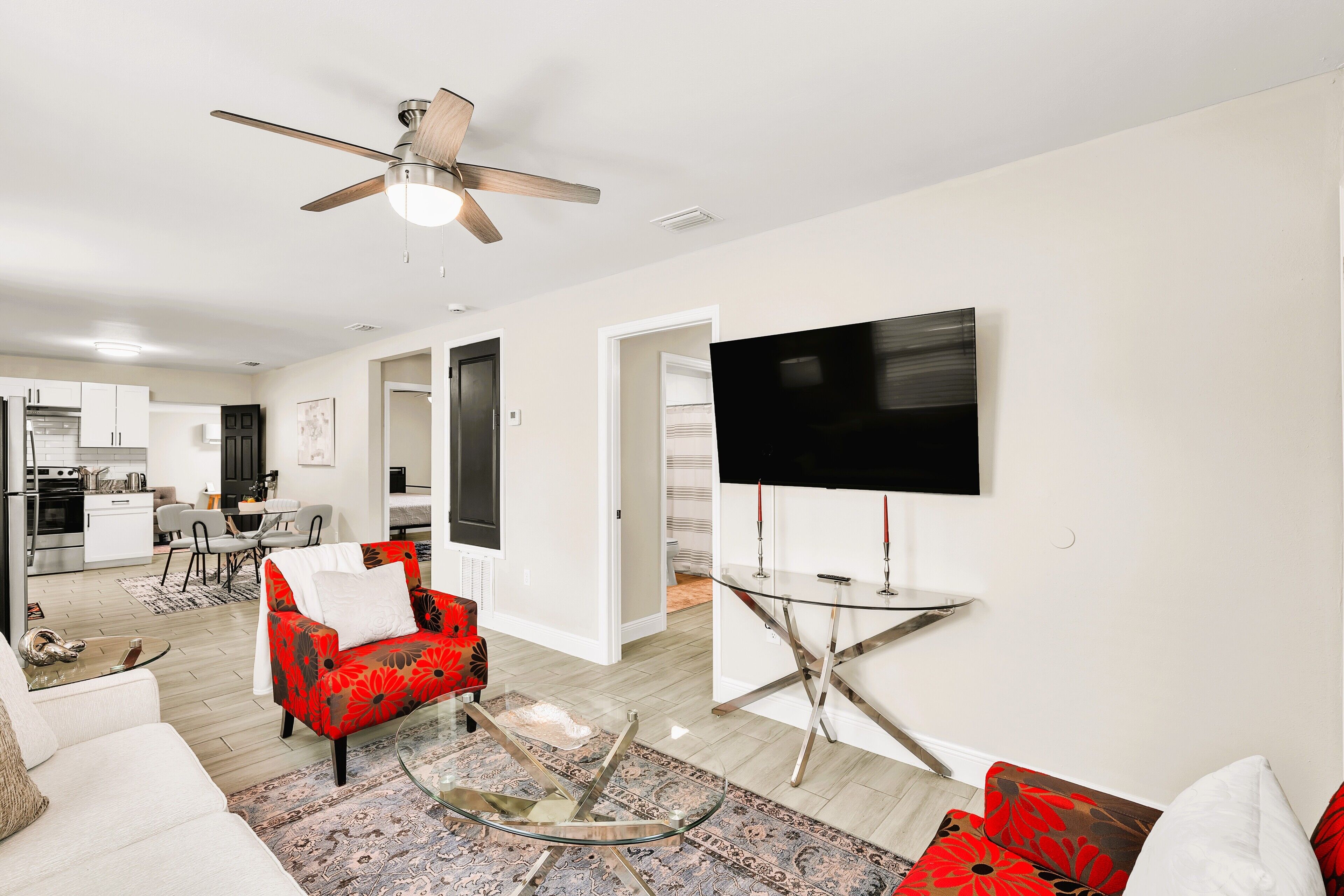 A modern living room with a flat-screen TV, a ceiling fan, and a dining area with a kitchen in the background.