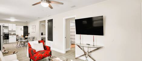 A modern living room with a flat-screen TV, a ceiling fan, and a dining area with a kitchen in the background.
