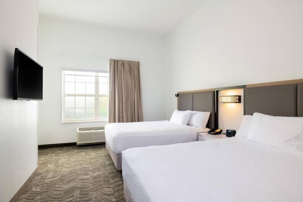 Une chambre d’hôtel avec deux lits, une télévision, une fenêtre avec des rideaux, une lampe fixée au mur et un thermostat.