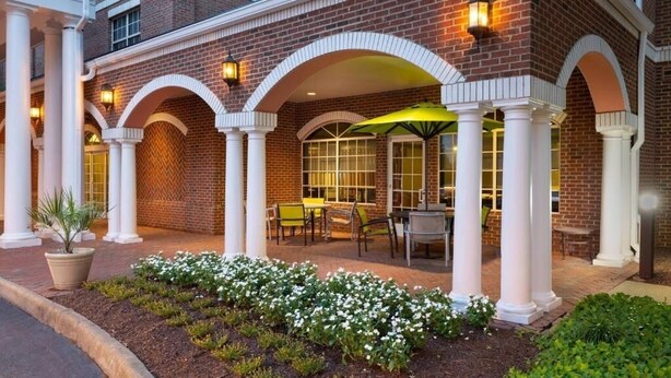 A brick building with white columns, outdoor seating, and a flower bed. - SpringHill Suites | Queen Suite | Outlets, Winery, Merchants Sq (Williamsburg)