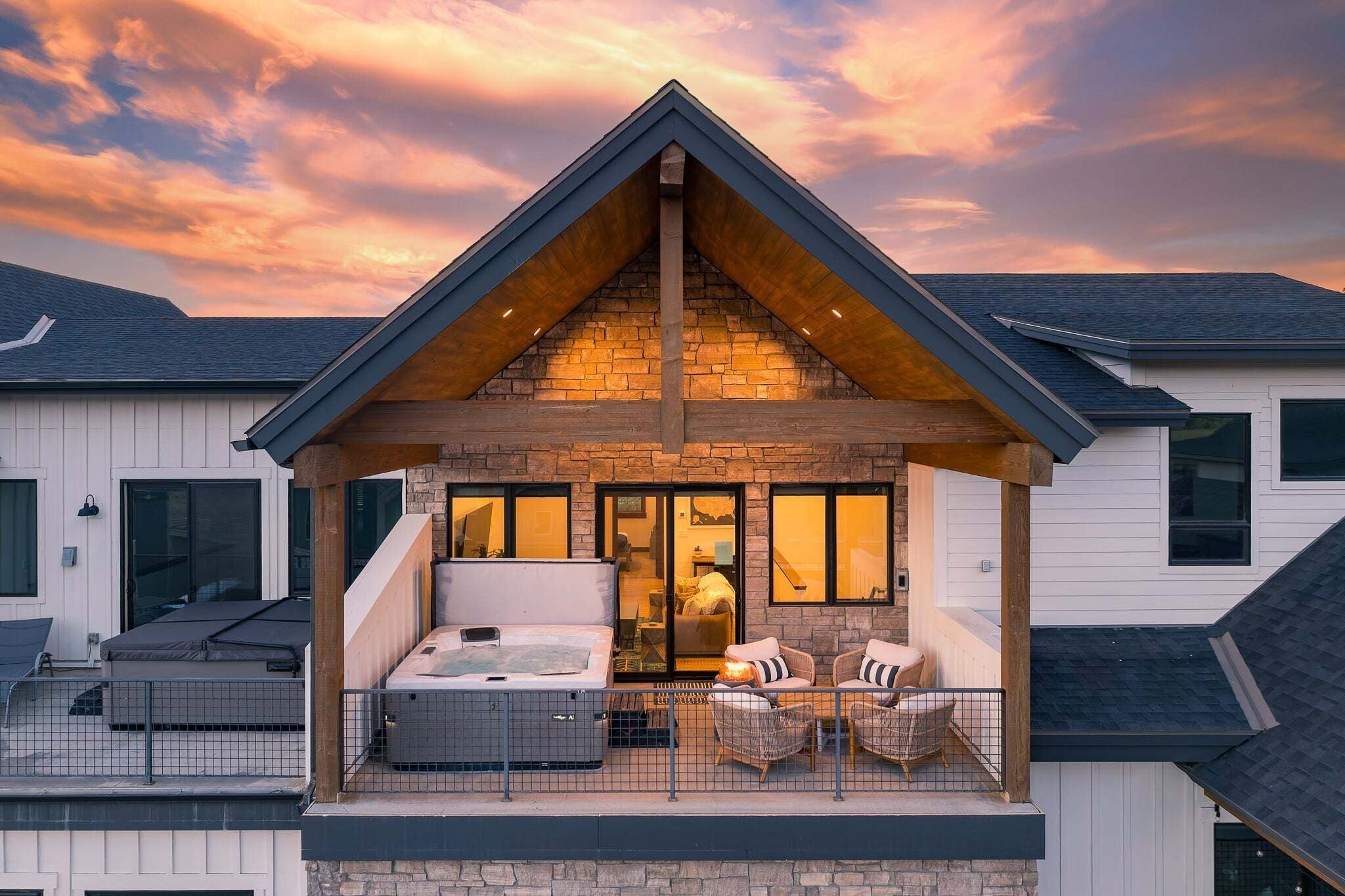 Une maison moderne avec une façade en pierre, un toit en bois et un balcon doté d’un espace salon extérieur et d’un jacuzzi.