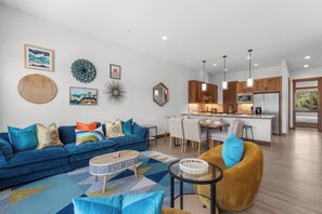 Ein modernes Wohnzimmer mit einer Couch, einem Couchtisch und einem gelben Sessel. Im Hintergrund ist eine Küche mit Holzschränken und ein Essbereich mit Stühlen erkennbar.