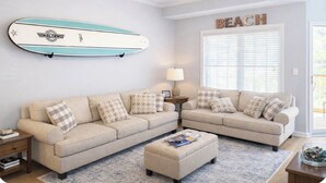 Una sala de estar con un sofá color crema, una mesa de centro, una tabla de surf colgada en la pared, una lámpara y una ventana con persianas.
