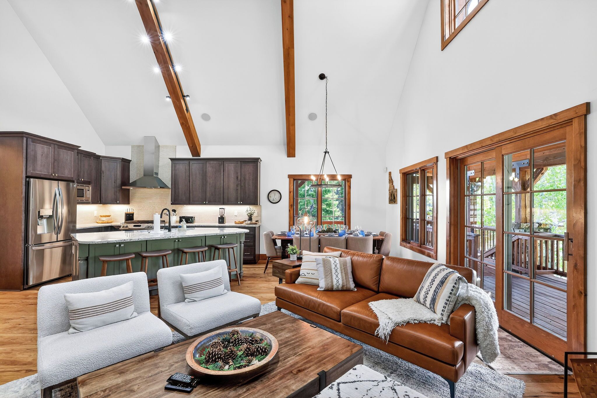 A spacious living area with a large brown leather sofa, two white armchairs, a wooden coffee table, and a dining area with a kitchen in the background.