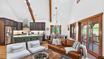 A spacious living area with a large brown leather sofa, two white armchairs, a wooden coffee table, and a dining area with a kitchen in the background.
