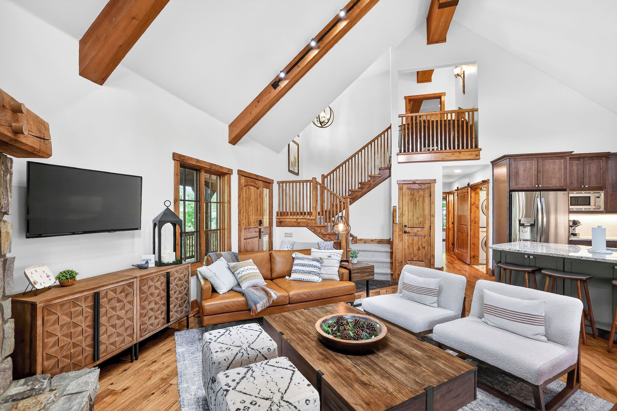 A spacious living room with a large TV, wooden furniture, a staircase, and a kitchen area.