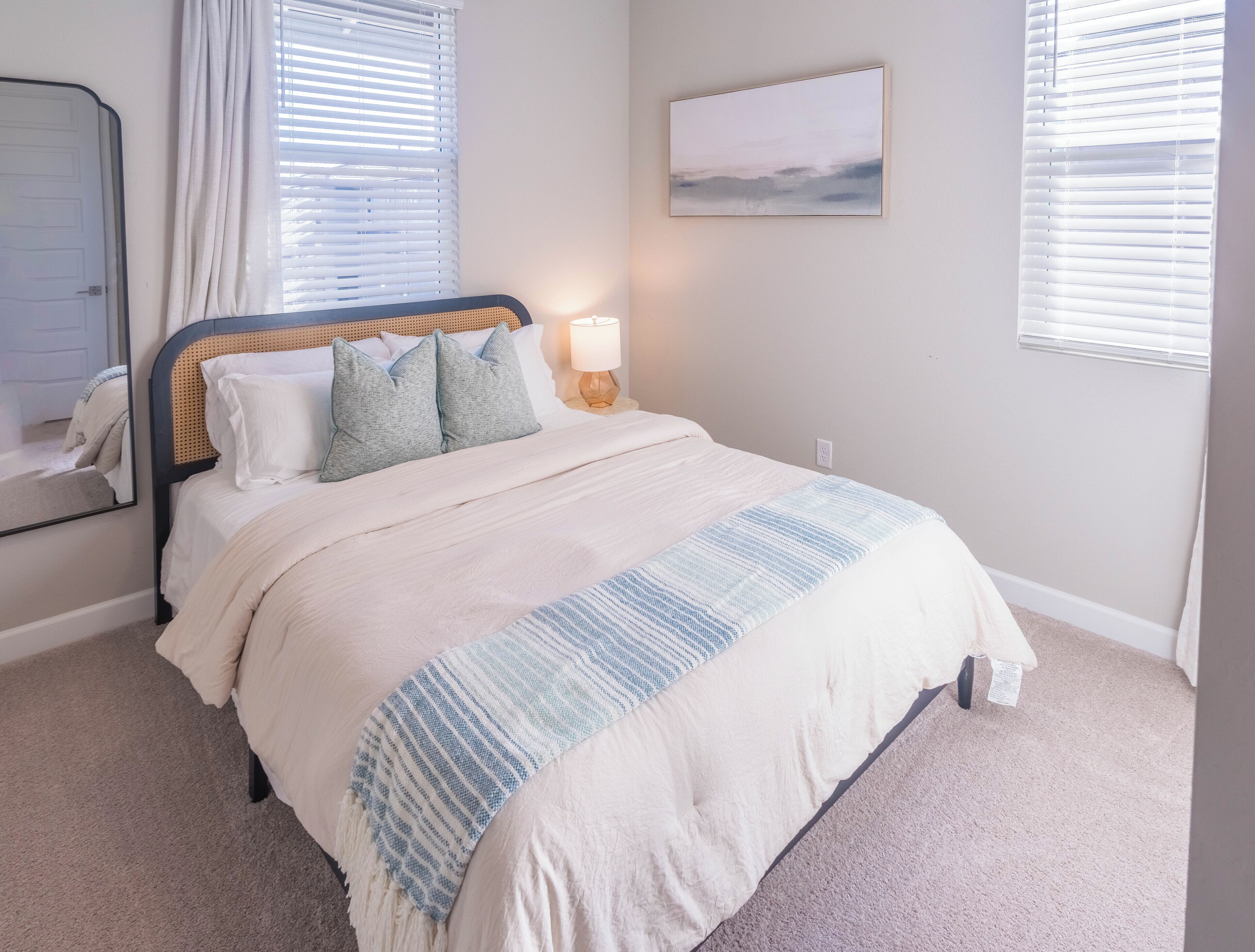 A bedroom with a bed, a bedside lamp, a window with blinds, a wall-mounted painting, and a door.