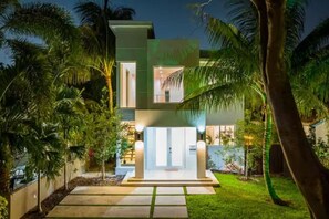 A modern two-story house with a well-lit entrance, surrounded by palm trees and a paved driveway. - 2020 Built Modern Luxe Home Heated Pool & Jacuzzi (Hollywood)