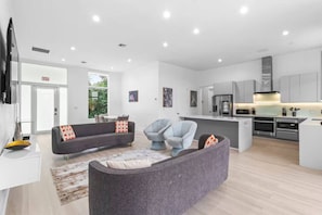 A modern living room with a grey sofa, a blue armchair, and a small coffee table. There is a kitchen area with grey cabinets and stainless steel appliances. - Modern Luxe 2021Built Heated Pool/Jacuzzi/Gameroom (Fort Lauderdale)