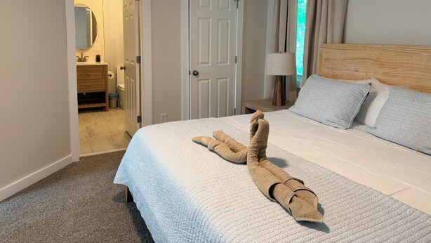 Ein Hotelzimmer mit Bett, Nachttisch, Lampe, einem Fenster mit Vorhängen und einem durchgegangene Tür sichtbaren Badezimmer.