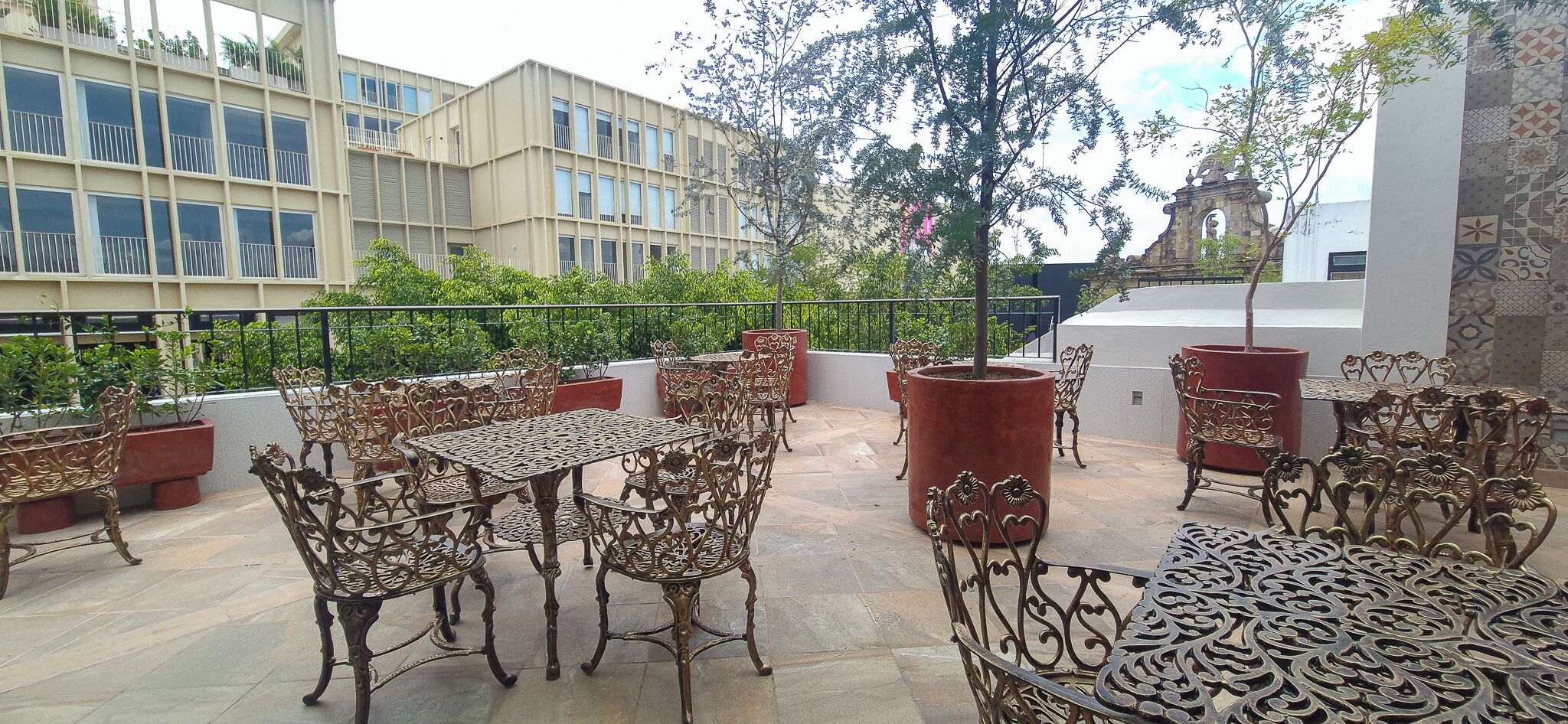 A rooftop patio with ornate metal furniture, a view of a building with large windows, and a decorative stone structure in the background.