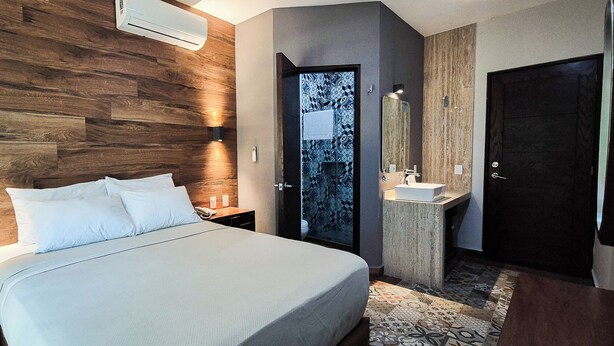 A modern hotel room with a wooden headboard, a bed with white linens, a bedside table, a built-in vanity with a mirror, and a door leading to a bathroom. - Gran Vila Nai Boutique Hotel (Zapopan)