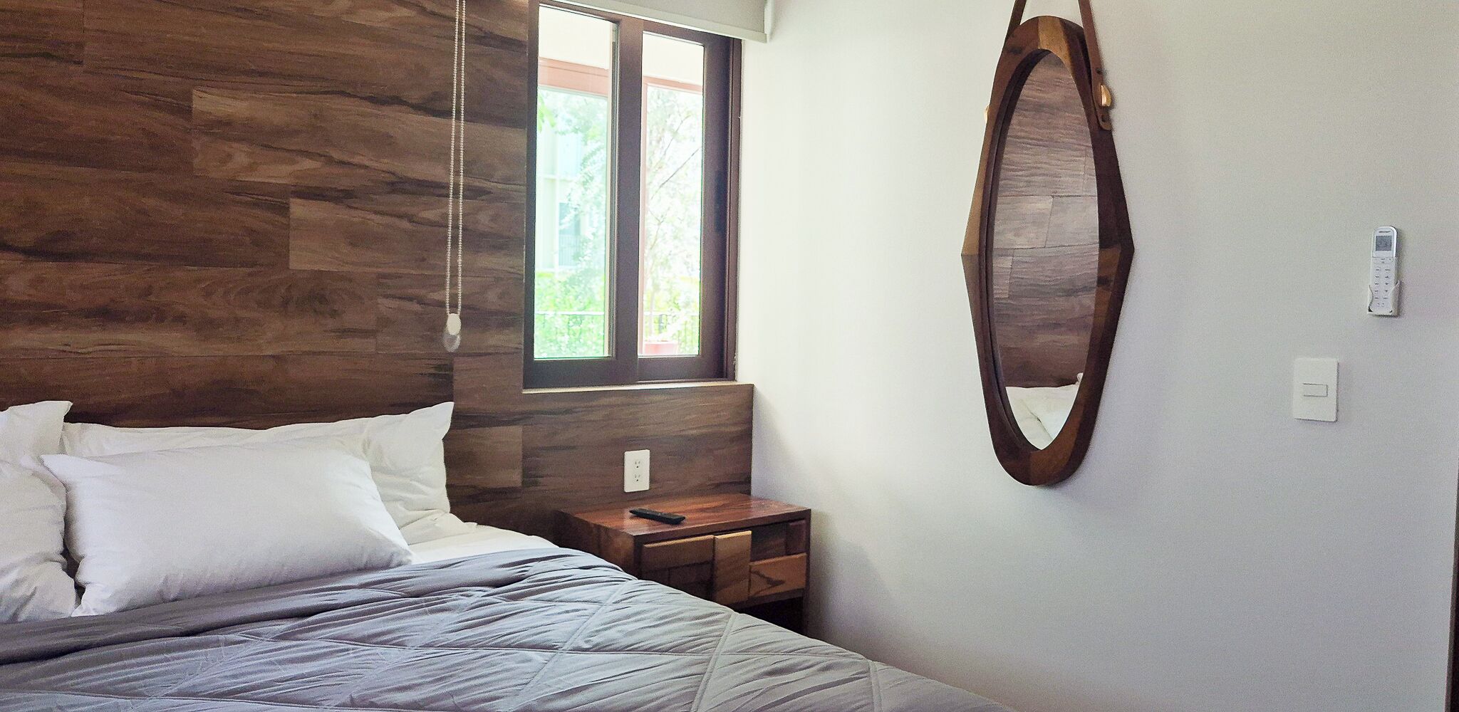 Un dormitorio con cabecera de madera, cama con ropa de cama blanca, mesita de noche con control remoto, espejo montado en la pared y ventana con vistas a zonas verdes.