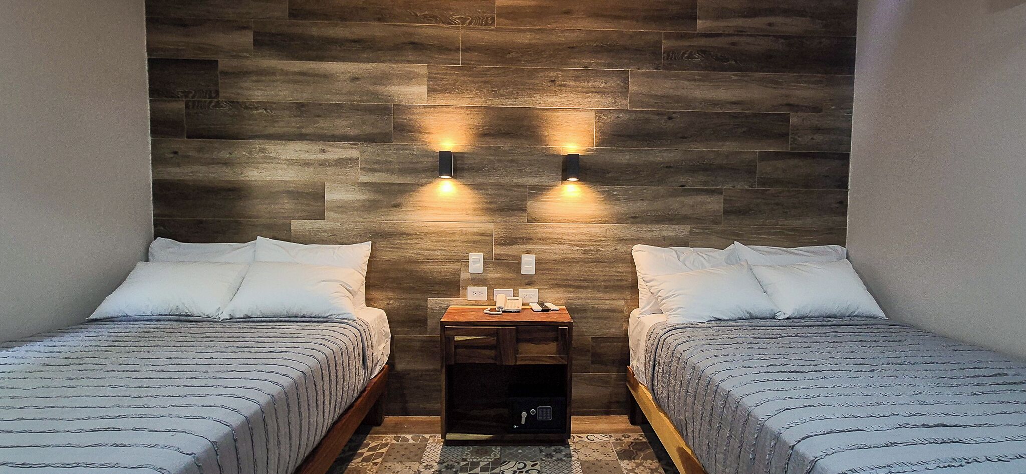 Two single beds with white linens, a wooden nightstand with a lamp, and a wall-mounted light fixture.