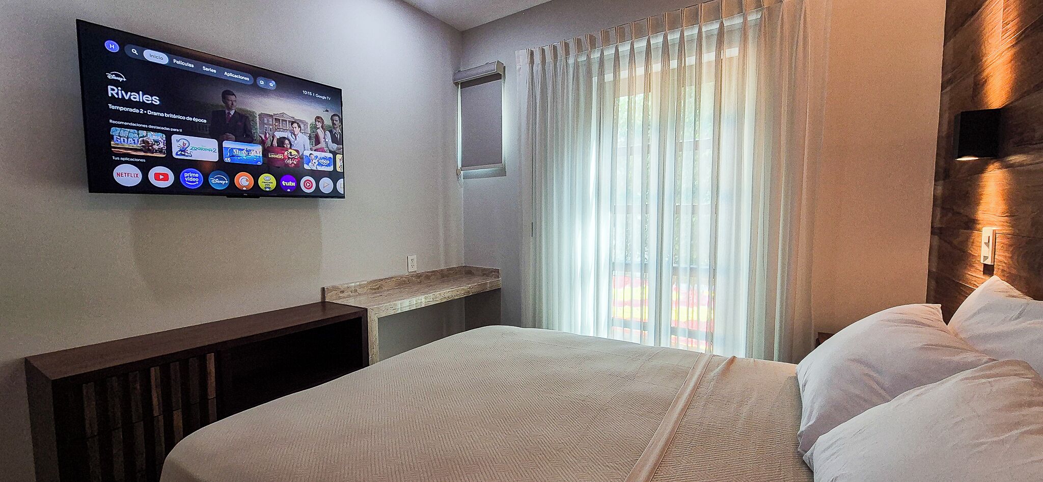 Ein Hotelzimmer mit Bett, einem an der Wand befestigten Fernseher, einem Fenster mit Vorhängen und einem Nachttisch.