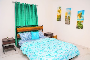 Ein Schlafzimmer mit einem hölzernen Bett, grünen Vorhängen, einer blumigen, blauen Bettdecke, zwei Nachttischen und drei gerahmten Bildern an der Wand.