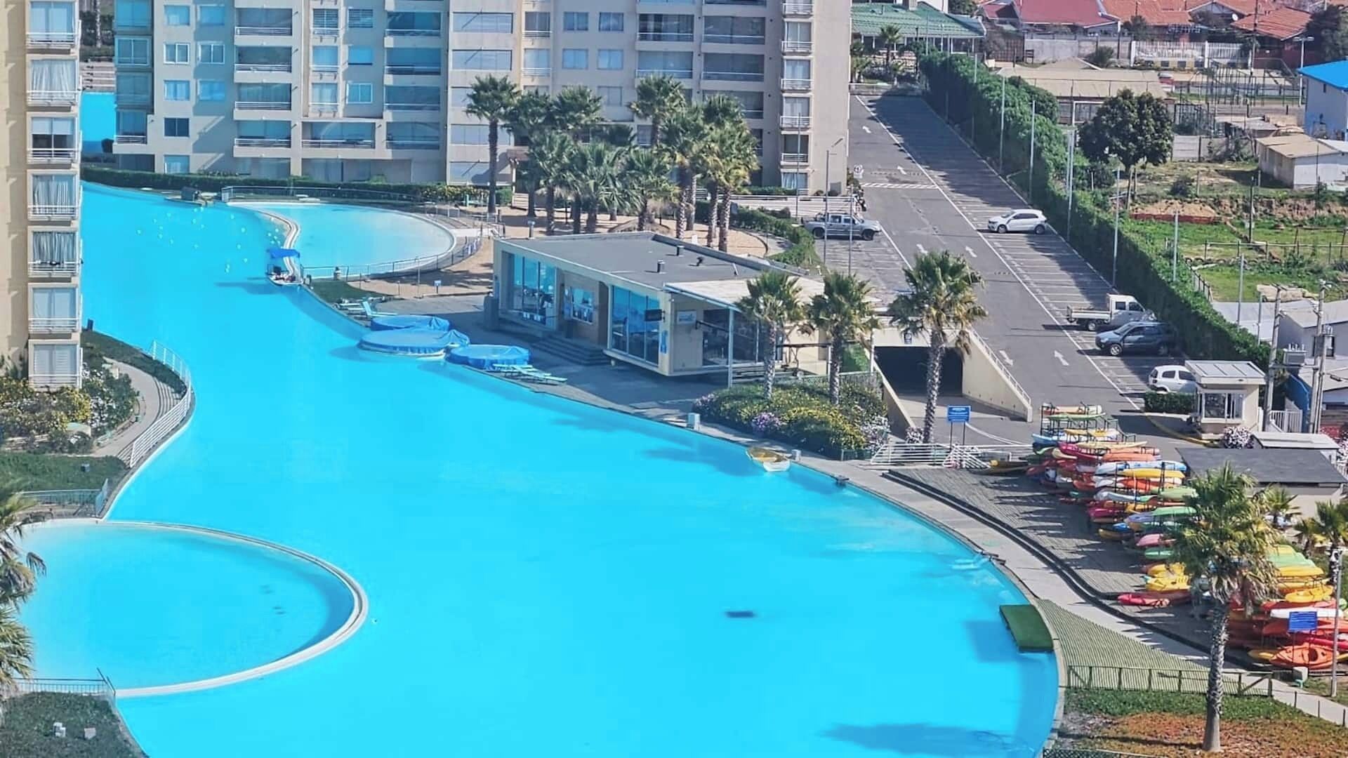A large swimming pool with a curved design, surrounded by palm trees and a parking area with colorful kayaks.