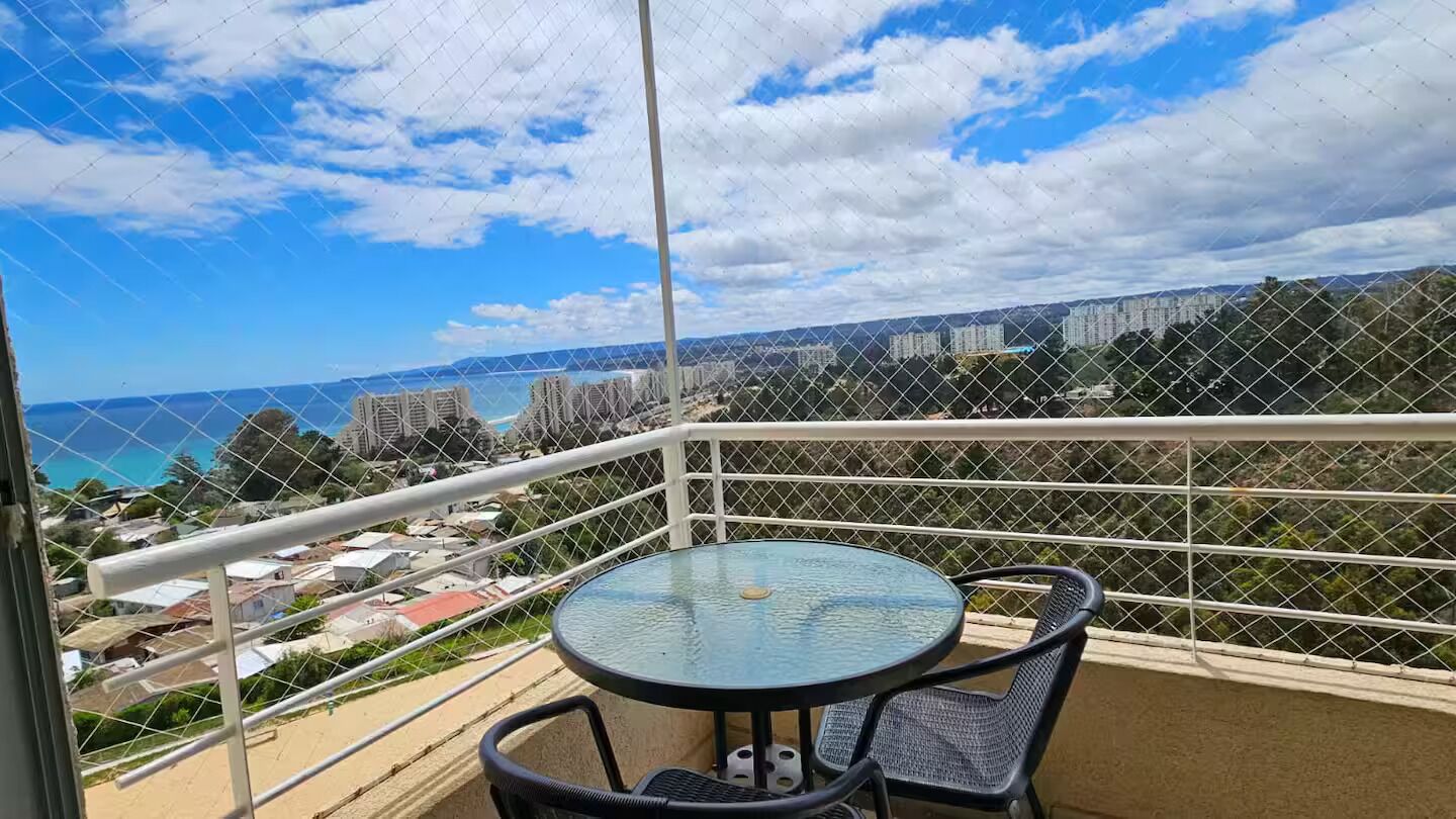 Un balcón con una mesa redonda de vidrio y sillas, con vista al mar y edificios, y zonas verdes.