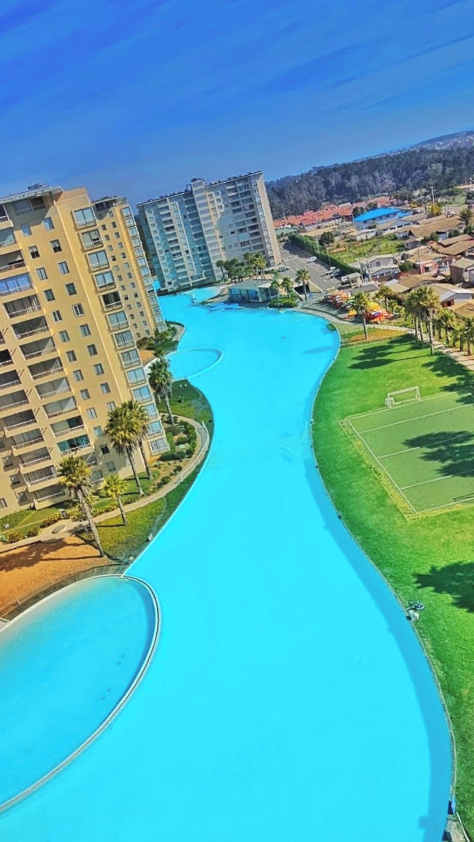 A large swimming pool with a curved design, surrounded by high-rise buildings and a grassy area with a soccer field.
