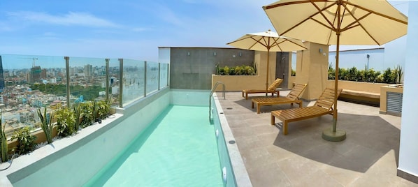 A rooftop pool with clear glass railings, wooden lounge chairs, and umbrellas.