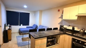 Een compacte keuken met een ingebouwde kookplaat, een eethoek met een tafel en stoelen, en een blauwe bank in de woonkamer.