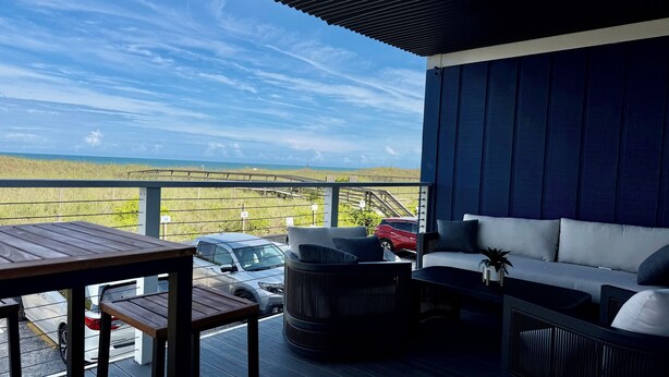 Un balcone con vista sulla spiaggia, un divano, un tavolo e un'auto parcheggiata all'esterno.