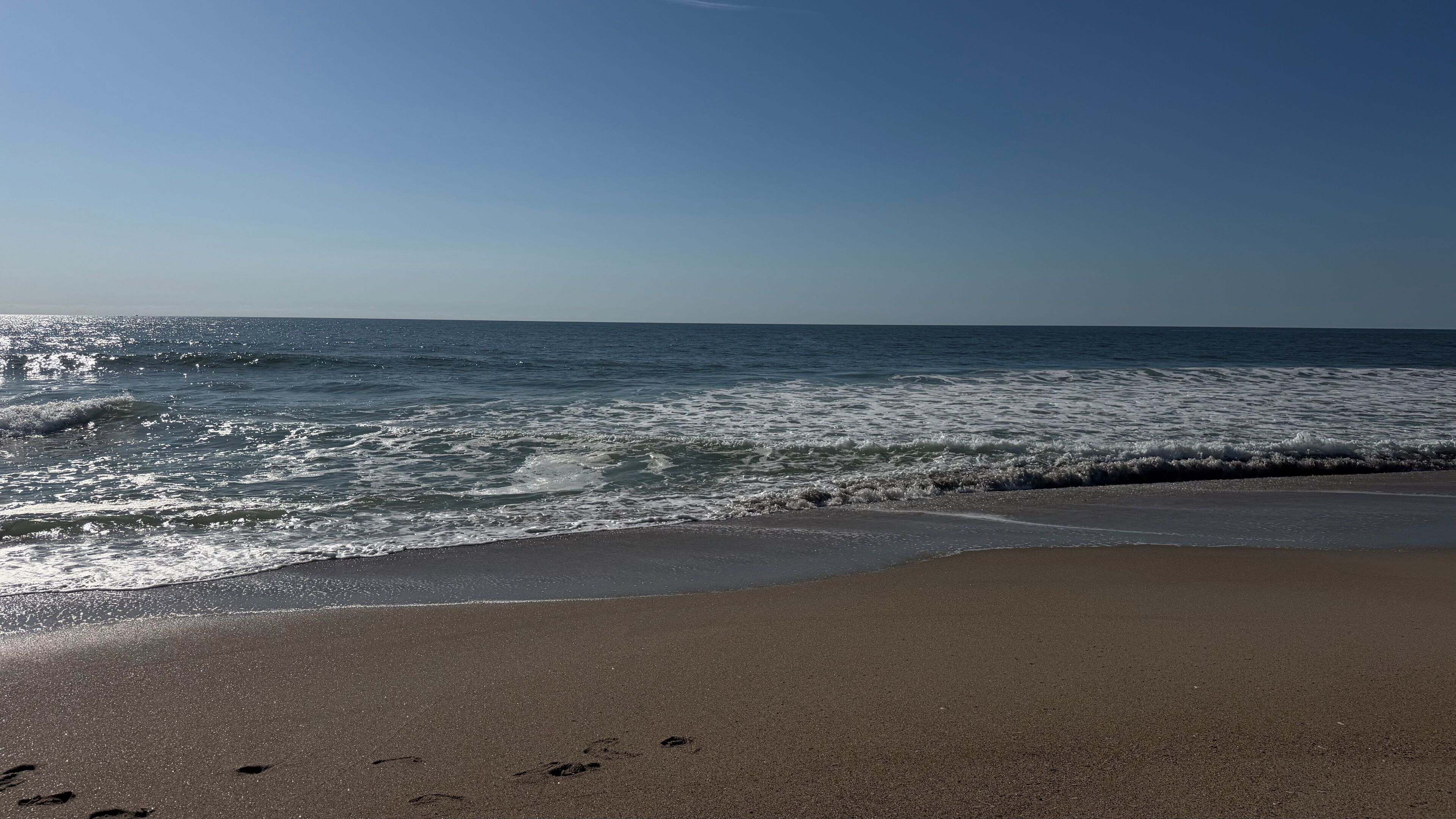 Una playa con huellas que se adentran en el agua, cielo despejado y olas tranquilas.