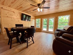 Ein Holz-Blockhaus-Innenraum mit Esstisch, Stühlen, einem an der Wand montierten Fernseher und einem Deckenventilator.
