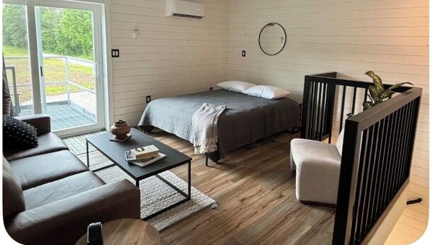 Una habitación con cama, sofá, mesa de centro, una mesita con un libro, espejo y ventana con vistas a la vegetación.