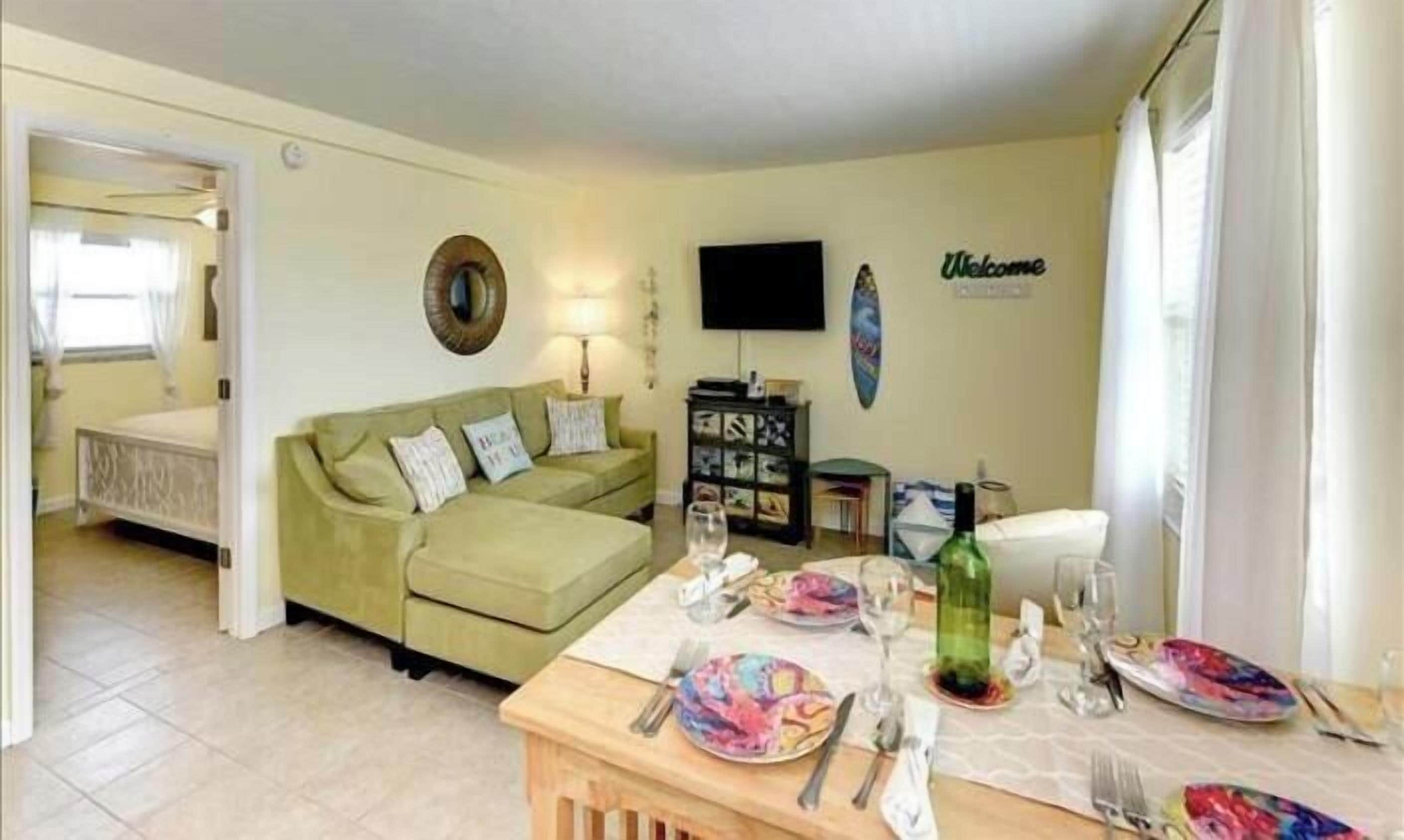 Ein Wohnzimmer mit einem grünen Sofa, einem Esstisch gedeckt mit Tellern und Besteck, einem Fernseher und einem Schild mit der Aufschrift 'Welcome' an der Wand.