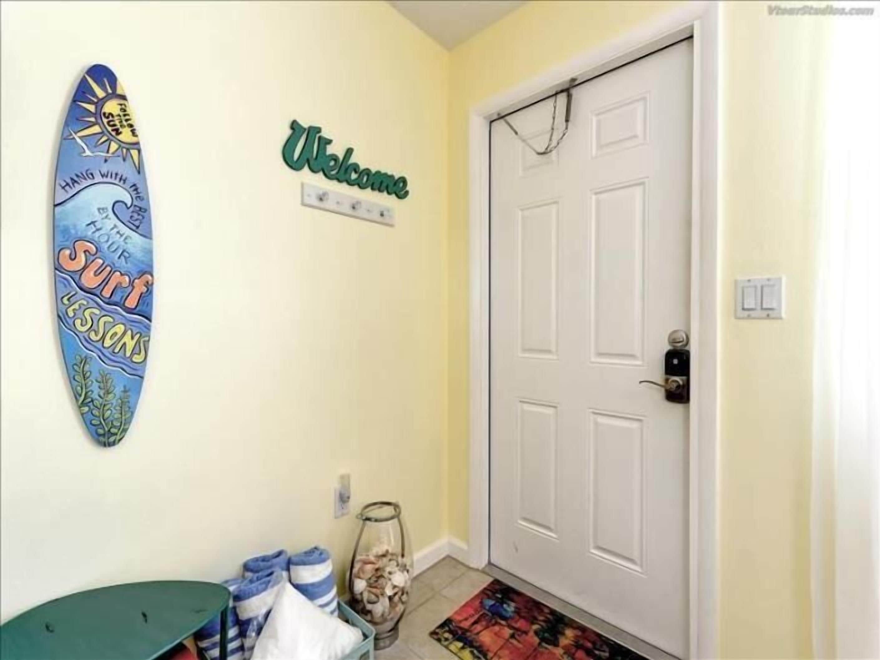 Een gang met een surfboard decoratie, een bord met de tekst welkom, een deur en een vloermat.