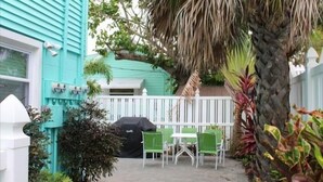 Une maison turquoise avec une clôture blanche, une terrasse avec des chaises vertes et une table, et un barbecue.