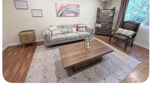 Uma sala de estar com sofá, mesa de centro, cadeira, luminária e janela.