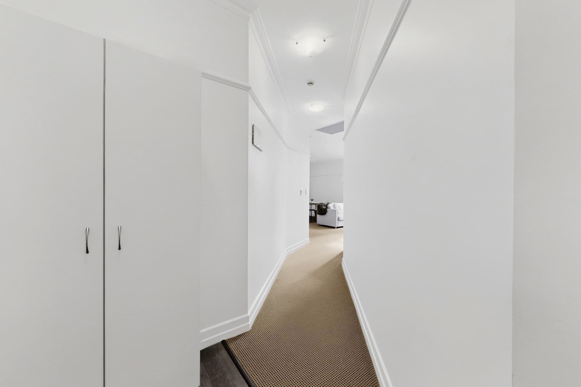 A narrow hallway with white walls and a carpeted floor leading to a room with a white sofa and a small table.