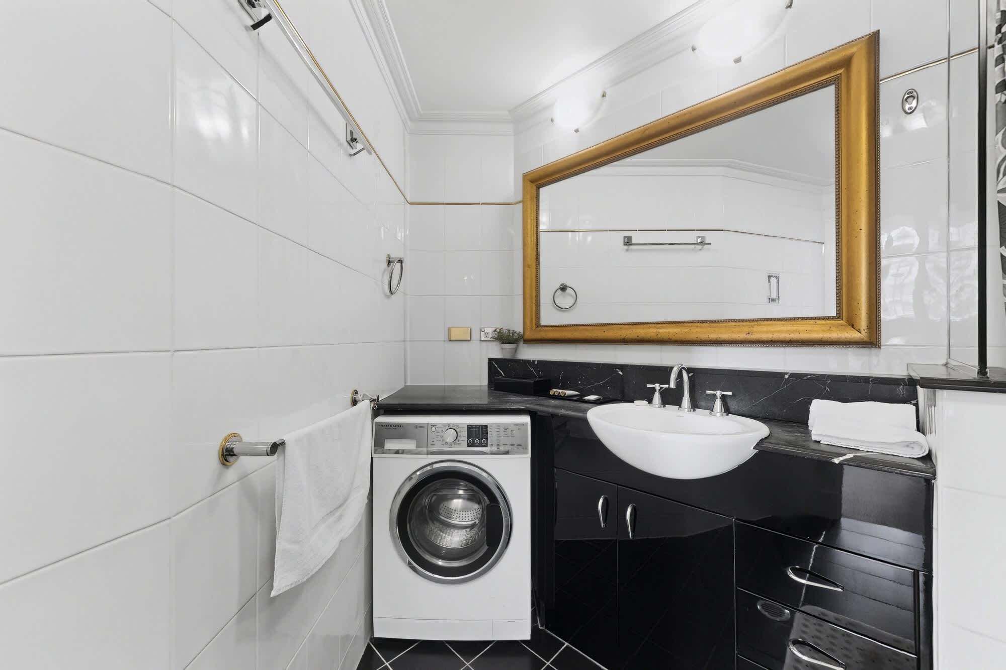 A compact bathroom with a washing machine, a sink, and a large mirror with a gold frame.