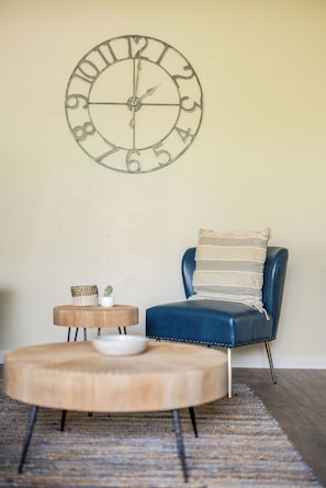 Una habitación con una mesa redonda de madera, una silla azul con un cojín a rayas y un reloj de pared.