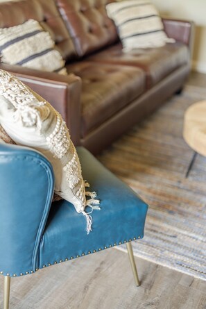 Un sillón azul con flecos, un plaid estampado y un sofá de cuero marrón con cojines a rayas al fondo.