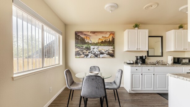 A modern kitchen with white cabinets, a dining area with a glass table and grey chairs, and a large window with blinds.