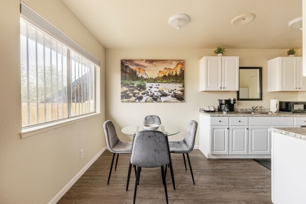 A modern kitchen with white cabinets, a dining area with a glass table and grey chairs, and a large window with blinds.