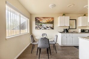 A modern kitchen with white cabinets, a dining area with a glass table and grey chairs, and a large window with blinds. - Cozy 1BR Near Fresno State - WiFi + AC (Fresno)