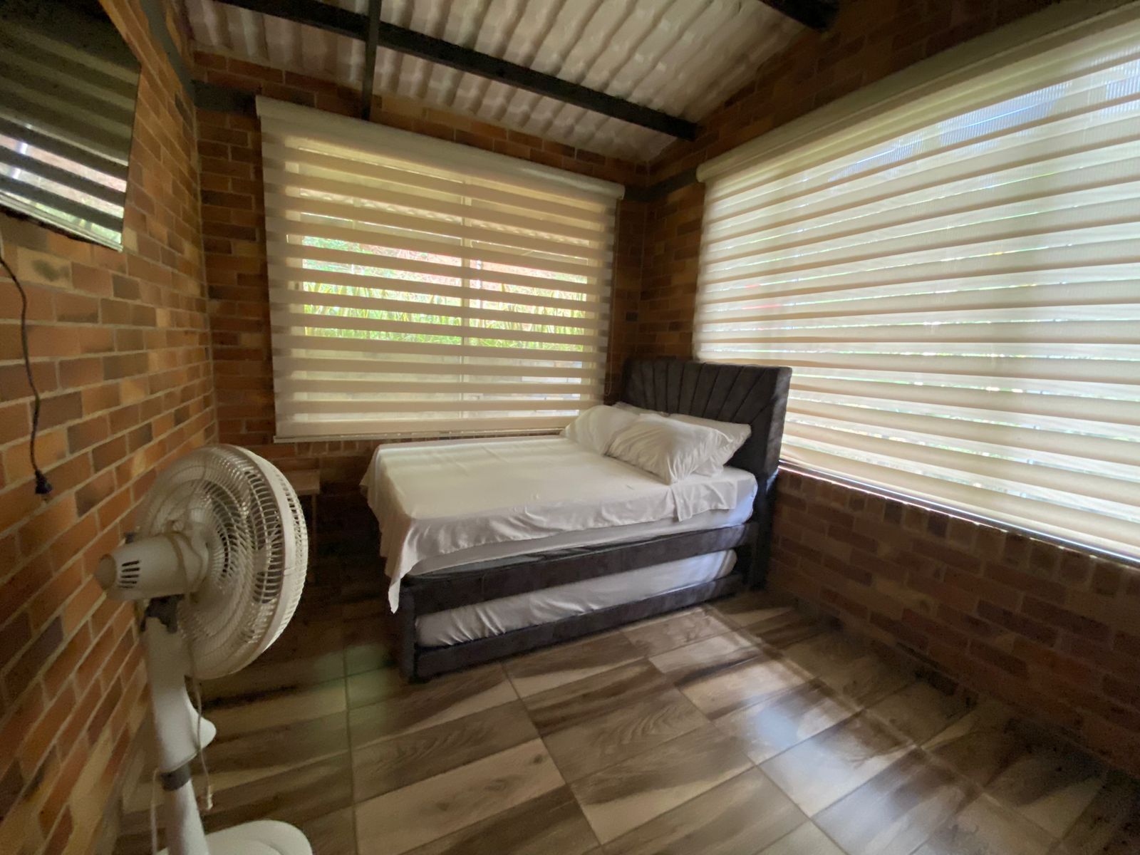 Una habitación con cama, ventilador y ventanas grandes con persianas.