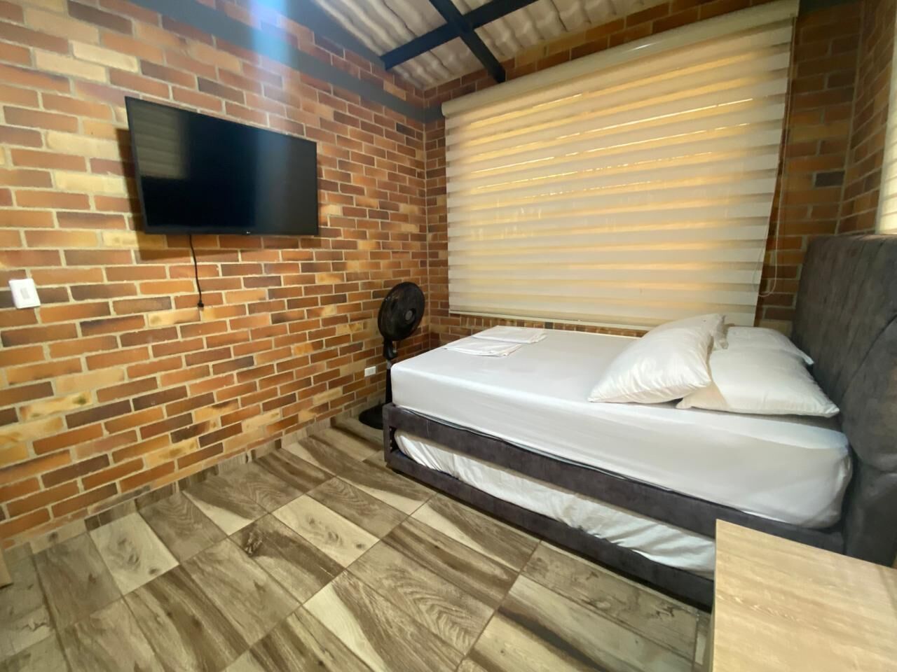 Een compacte kamer met een bakstenen muur, een flatscreen televisie, een bed met wit beddengoed, een ventilator en een raam met zonwering.