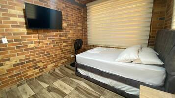 Een compacte kamer met een bakstenen muur, een flatscreen televisie, een bed met wit beddengoed, een ventilator en een raam met zonwering.
