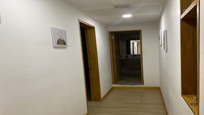 Un pasillo con suelo de madera, paredes blancas y techo con diseño de cuadrícula. Hay puertas a ambos lados y se ve un espejo a lo lejos.