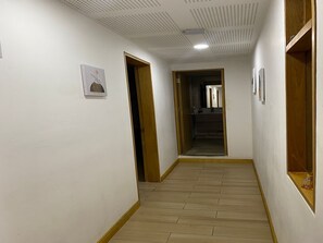 Ein Flur mit Holzboden, weißen Wänden und einer Decke mit Rasteroptik. Auf beiden Seiten befinden sich Türen, und in der Ferne ist ein Spiegel zu sehen.