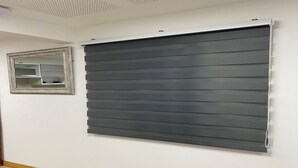 Habitación con una persiana enrollable grande de color oscuro, un espejo pequeño y suelo de madera.