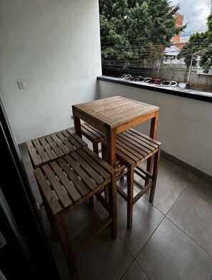 Ein Balkon mit Holztisch und Bank, ein Fenster mit Blick auf Bäume und parkende Autos.