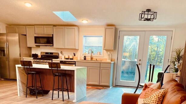 A modern kitchen with a central island, a dining area with a sofa and coffee table, and a view of the outdoors through large windows. - TRAVELING WORKERS 3 bdrm/sleeps 6-7 hin Spring Grove/Surry/Prince George (Spring Grove)